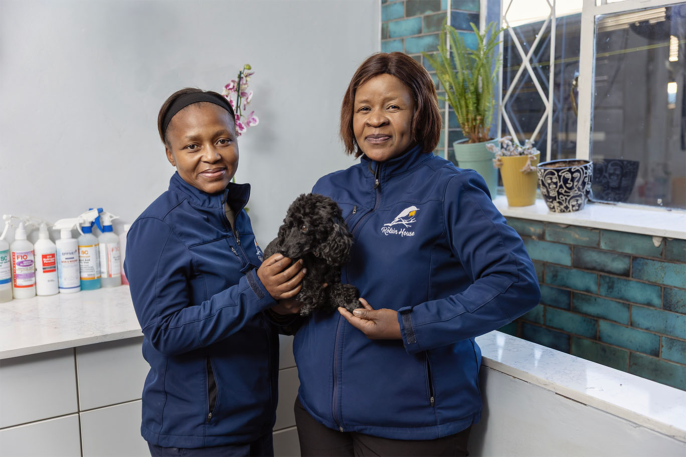 Head Pet Groomer Julia and Apprentice Mavis smiling warmly while holding a freshly groomed black poodle at the Johannesburg salon.